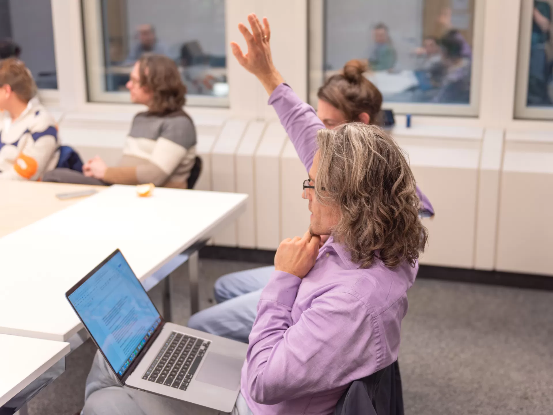 Eine Gruppe von Personen sitzt um einen Konferenztisch; ein Mann im Vordergrund hat einen aufgeschlagenen Laptop auf den Knien und hebt die rechte Hand für eine Wortmeldung.