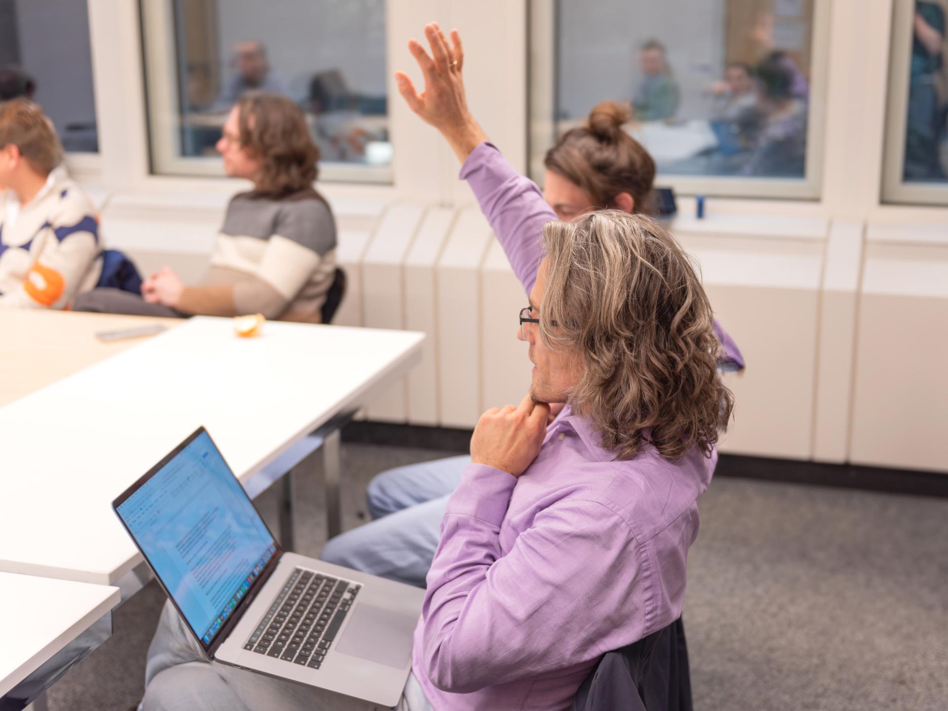 ... an der Hochschule diskutiert ... Eine Gruppe von Personen sitzt um einen Konferenztisch; ein Mann im Vordergrund hat einen aufgeschlagenen Laptop auf den Knien und hebt die rechte Hand für eine Wortmeldung.