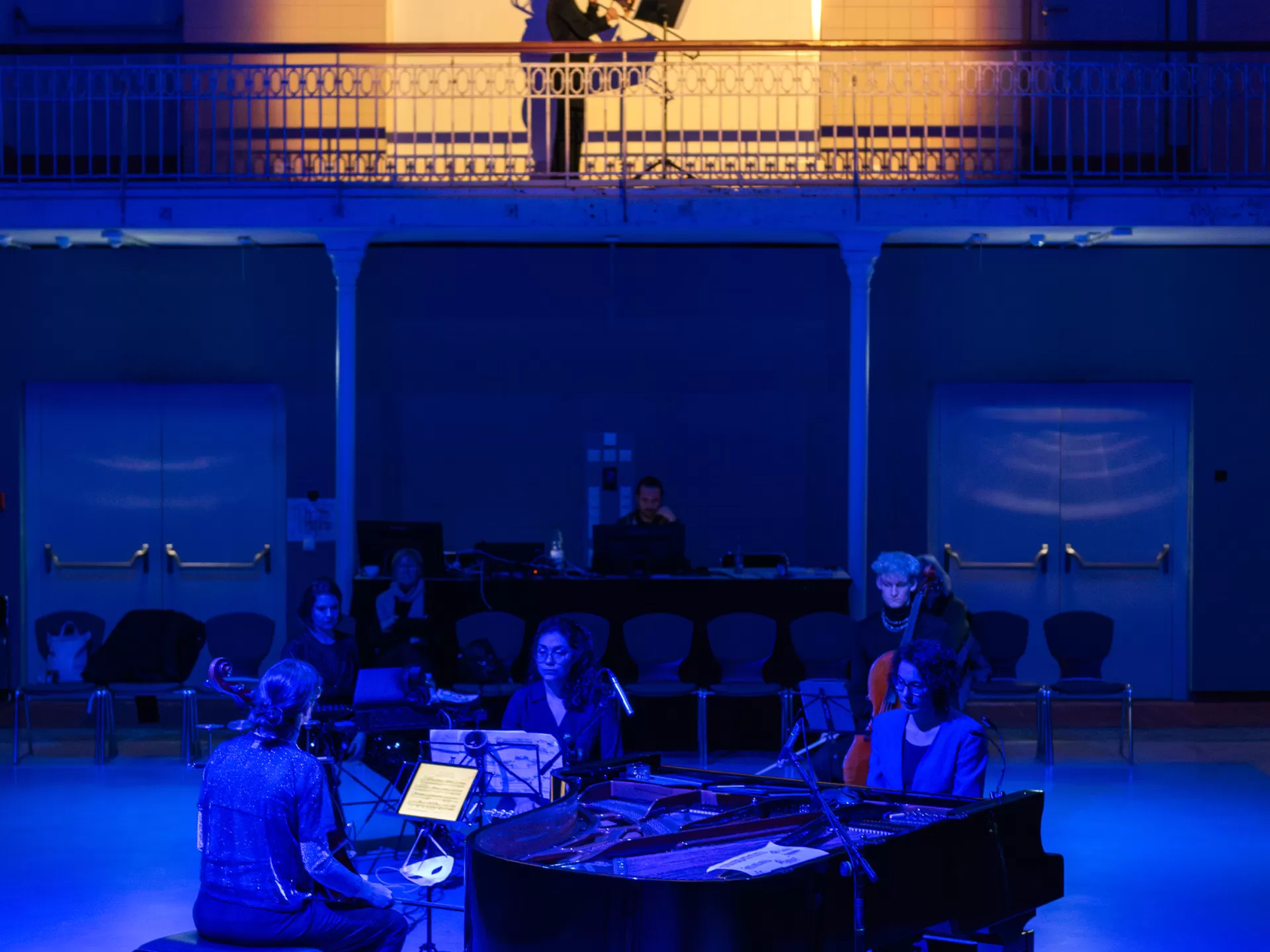Aufführungsfoto: Die Posaunistin spielt auf einer Empore in einem warmen Spot. Unten spielen die anderen Musiker*innen. Sie sind in blaues Licht gehüllt.