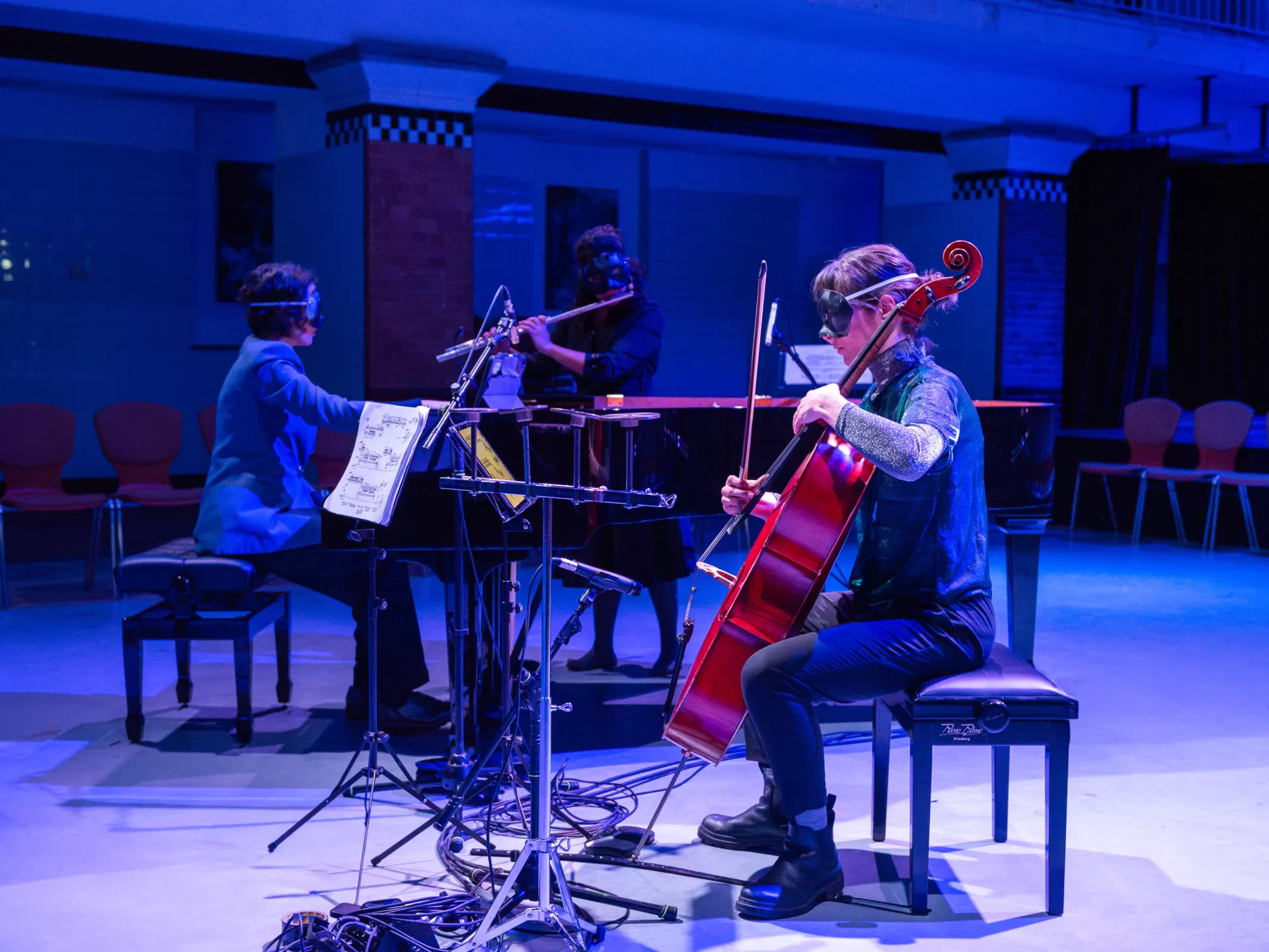 Eine Flötistin, eine Pianistin und eine Cellistin musizieren im Alten Halltenbad. Vor den Augen tragen sie schwarze Masken.