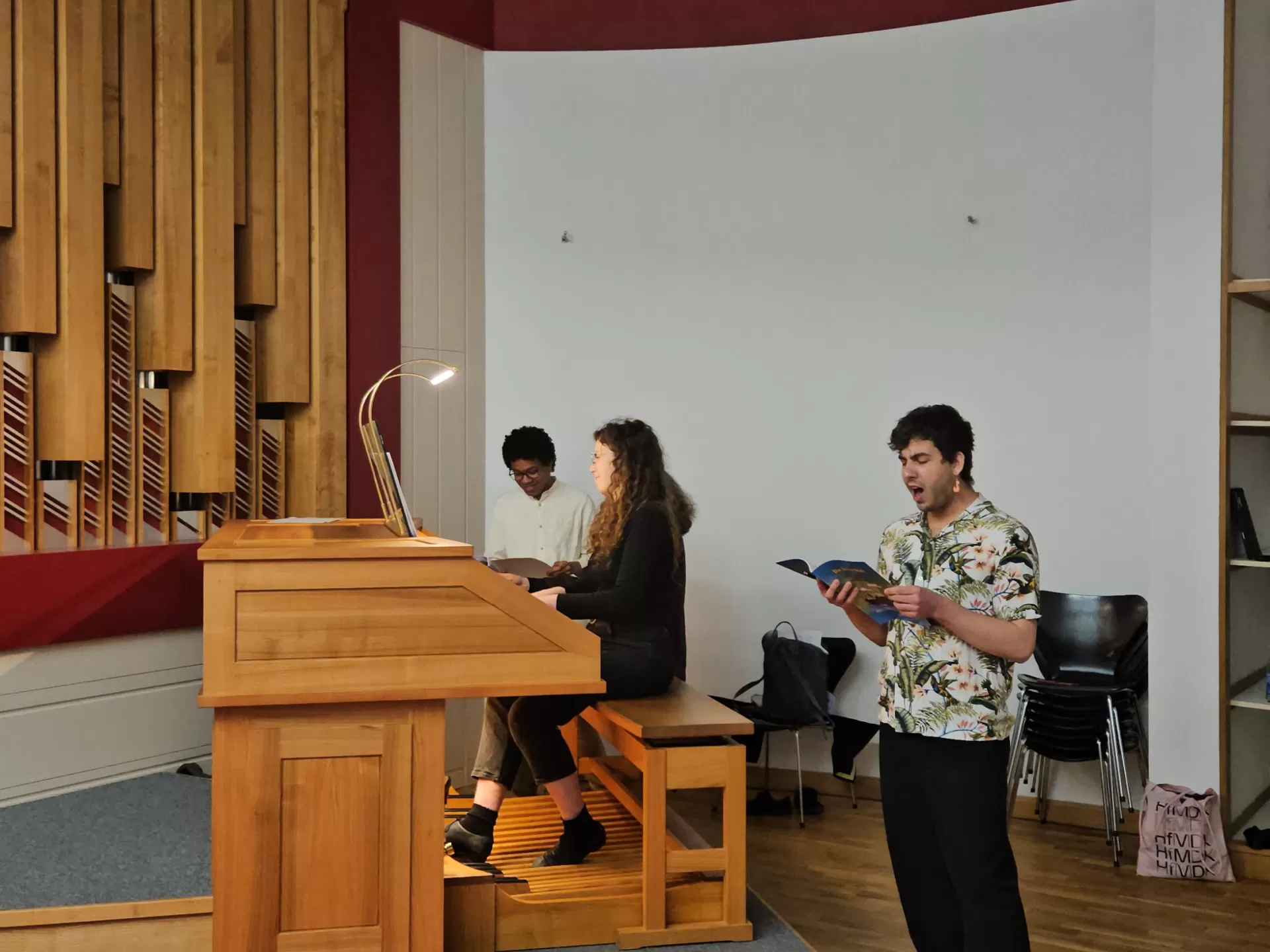 Blick in Orgelraum. Student liest laut energisch den Text, andere Studentin sitzt an der Orgel. Schulklasse hört zu.
