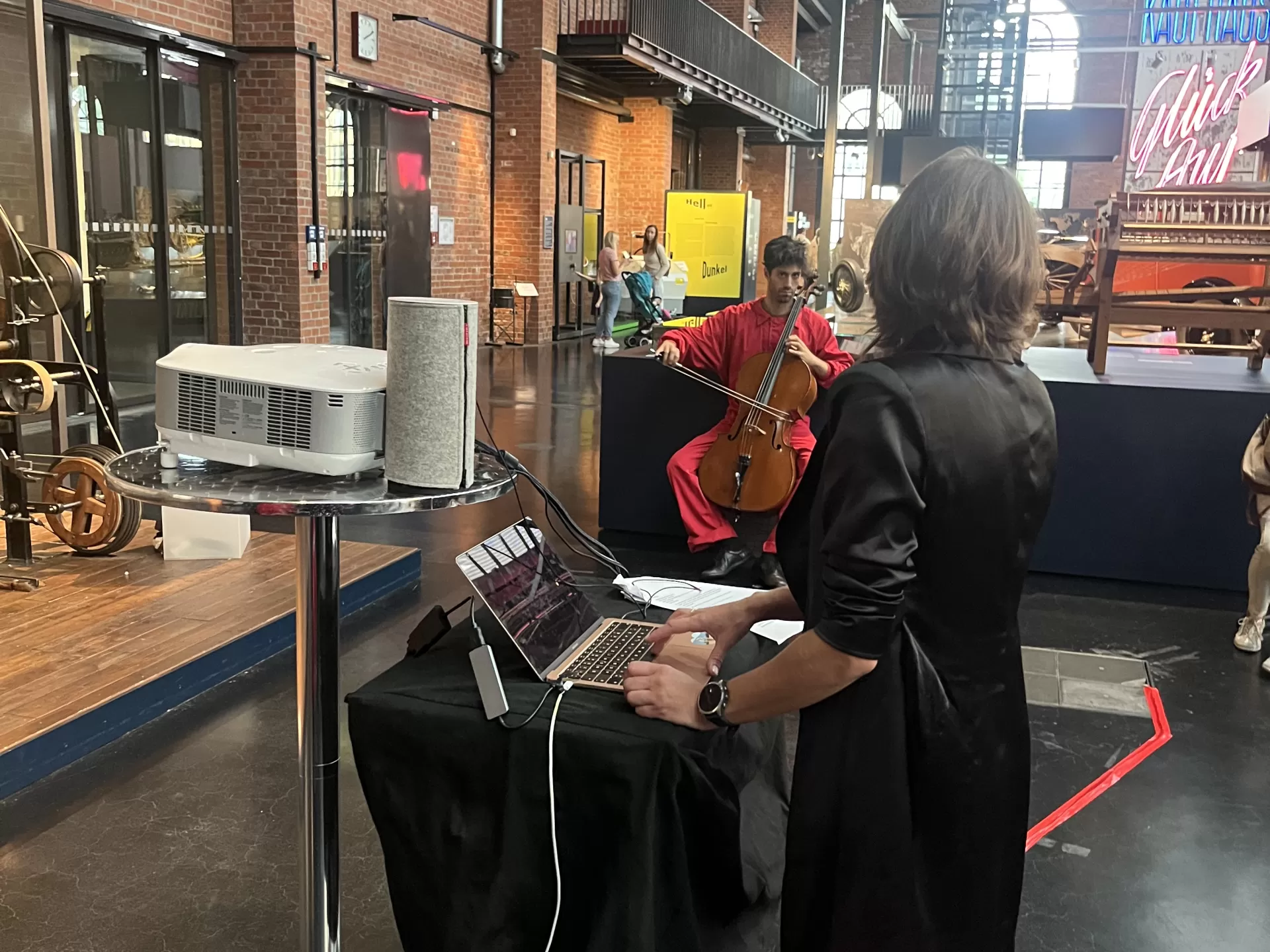 Rot-schwarz gekleidete Menschen musizieren im Industriemuseum. Im Vordergrund steht eine Studentin am Laptop.