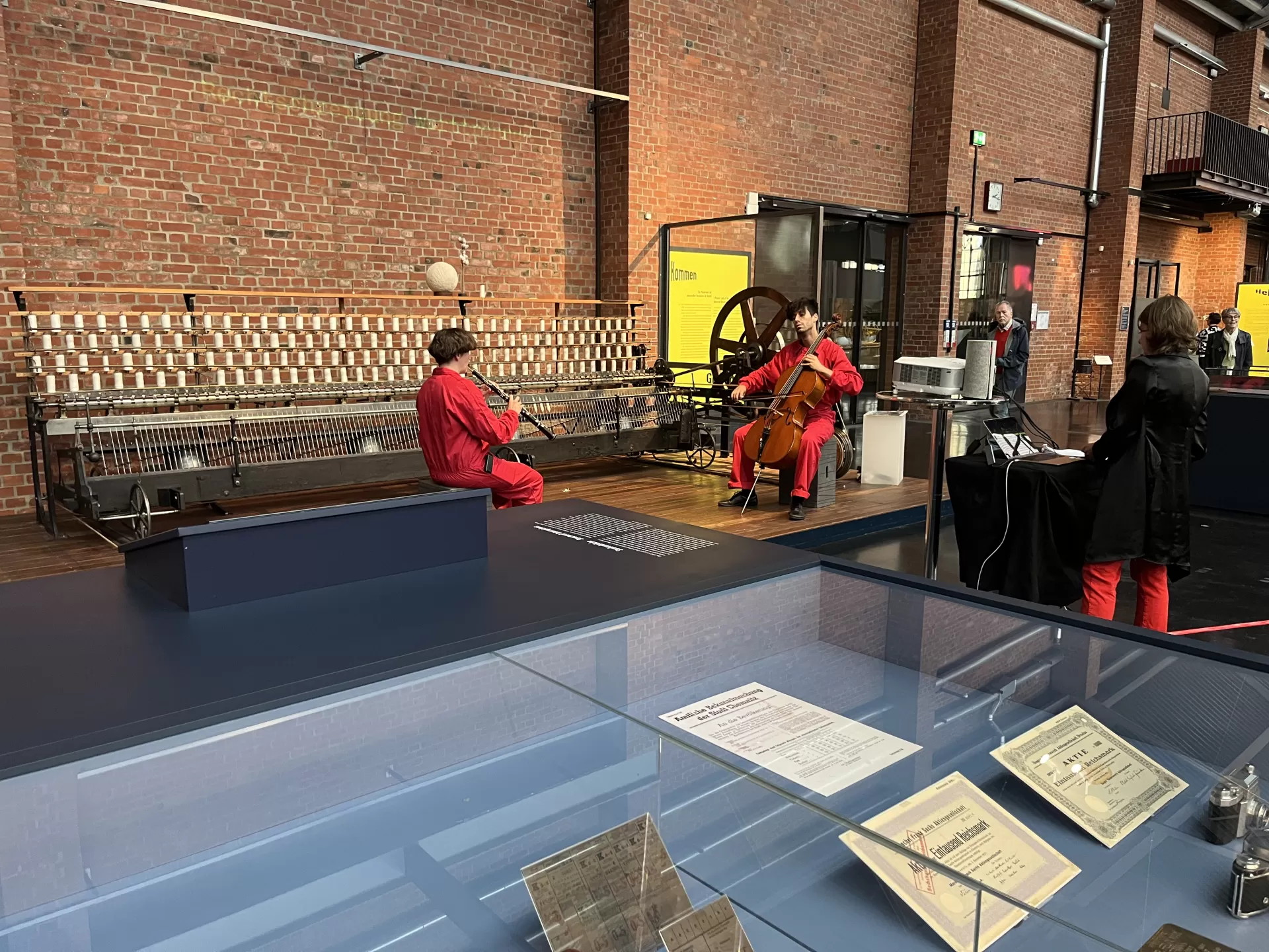 Rot-schwarz gekleidete Menschen musizieren im Industriemuseum. Zwei Personen vor einer Backsteinwand spielen Oboe und Cello. Davor steht eine Person am Laptop.