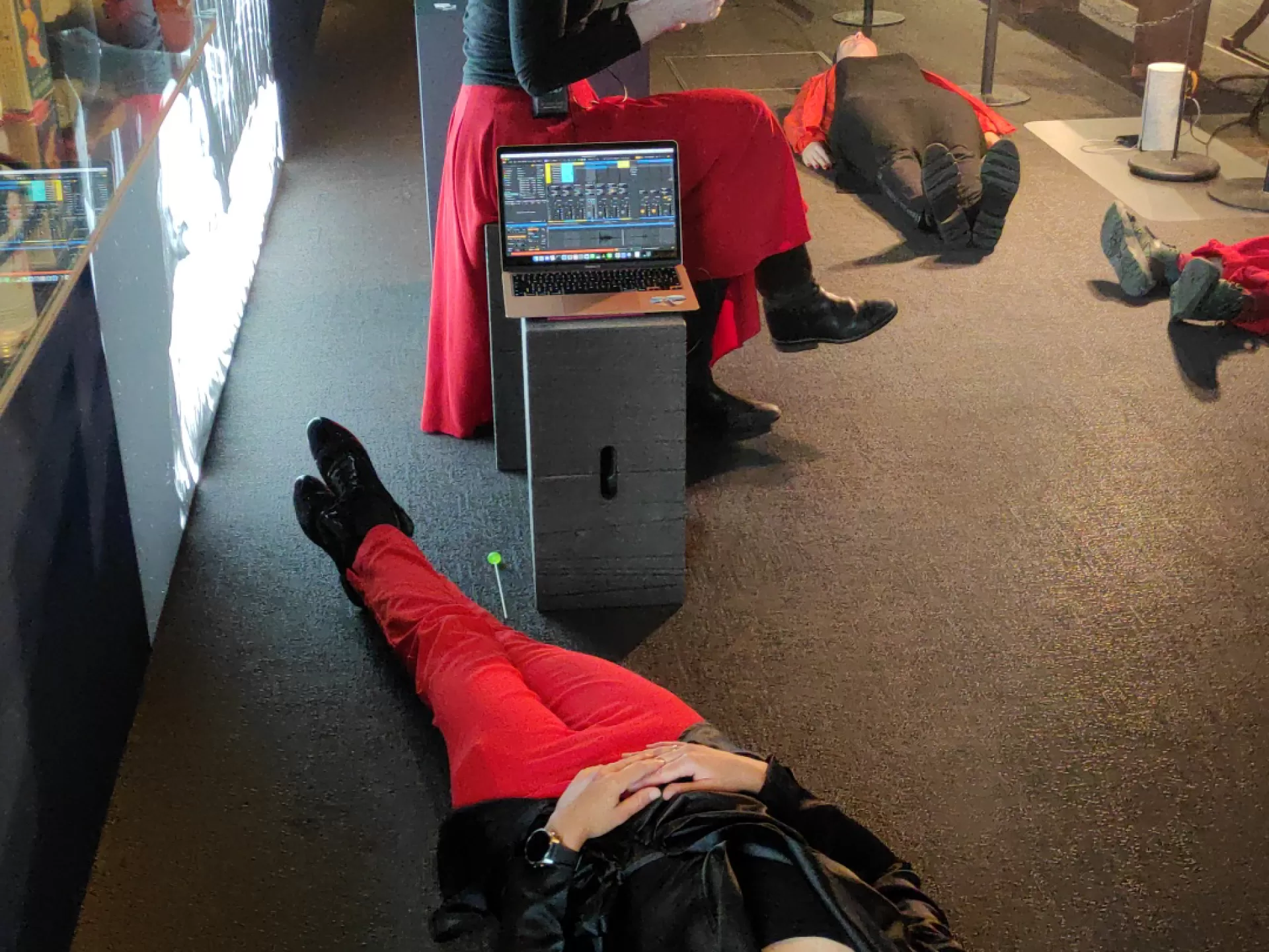 Eine Person sitzt inmitten einer Ausstellung und liest etwas vor. Weitere Personen liegen auf dem Boden und tragen Kopfhörer.