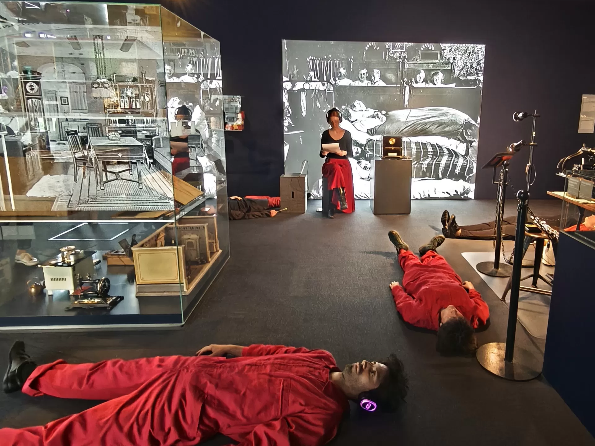 Mehrere Menschen in roten Overalls liegen mit Kopfhörern auf dem Boden in einer Ausstellung. Eine Person sitzt auf einem Klotz und liest etwas von einem Blatt Papier ab.