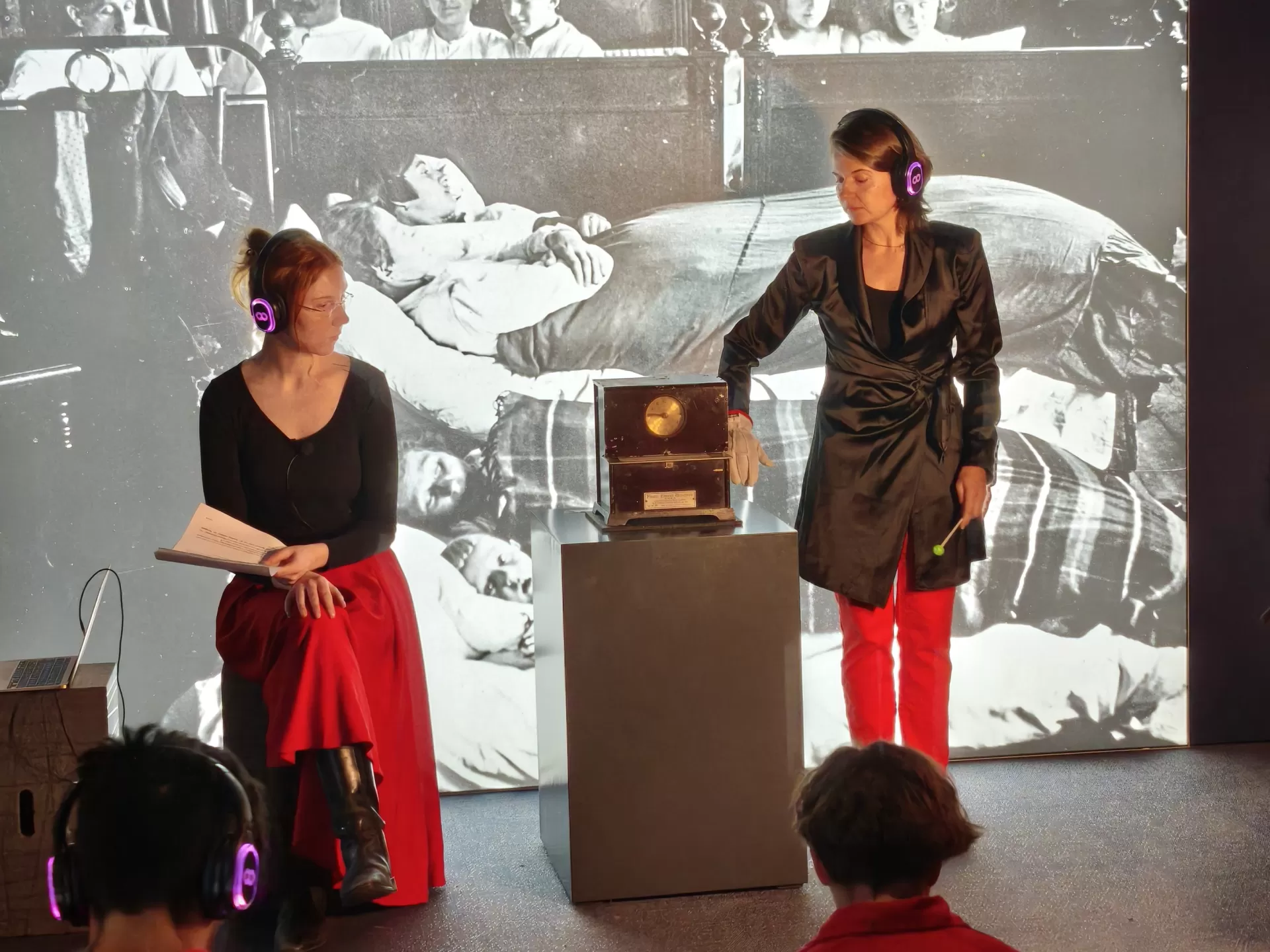 Rot-schwarz gekleidete Menschen mit Kopfhörern im Industriemuseum. Eine Person berührt eine antik aussehende Uhr.