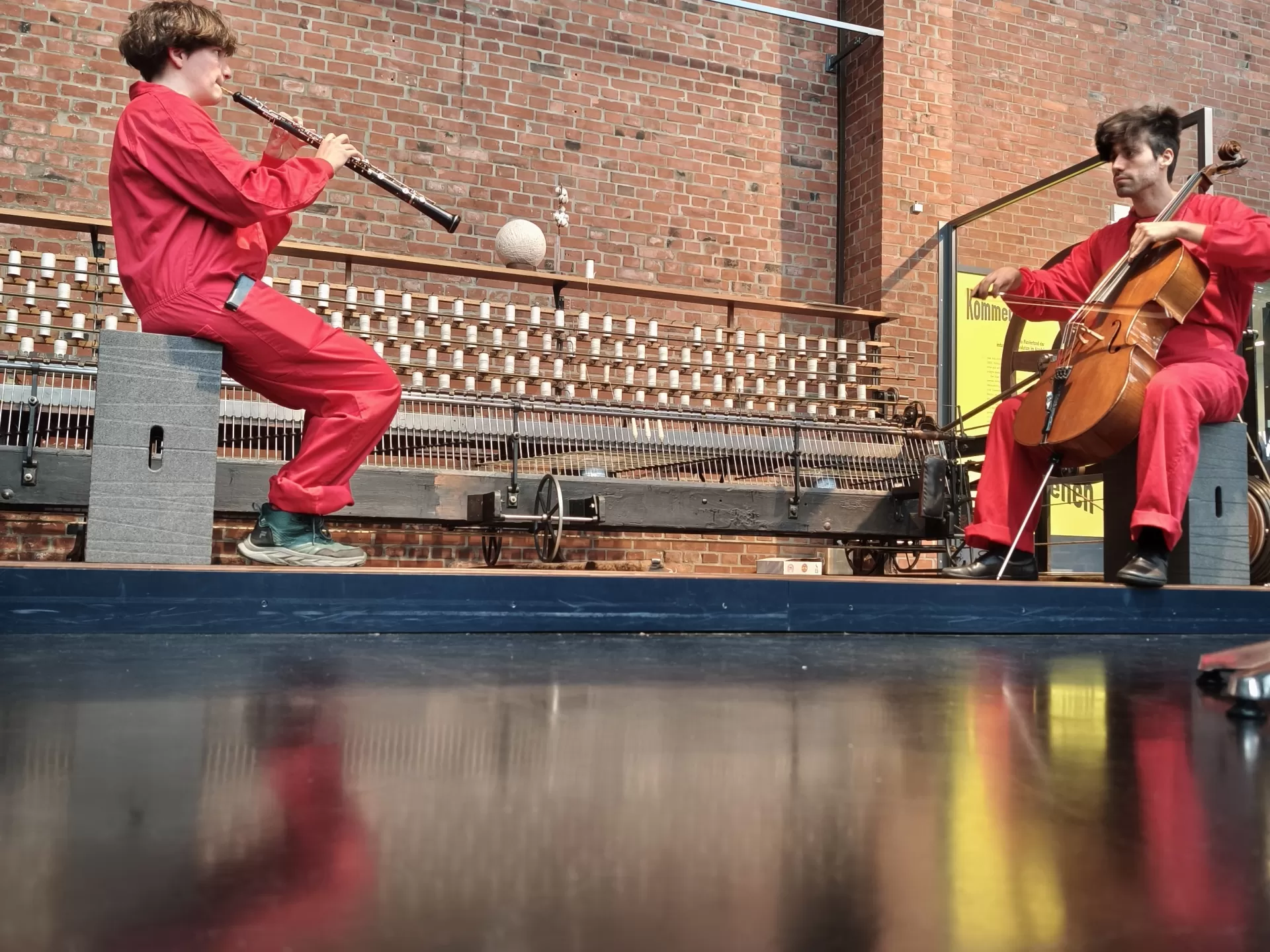 Zwei Studierende in roten Overalls sitzen vor einer Backsteinwand im Museum und spielen Oboe bzw. Cello.