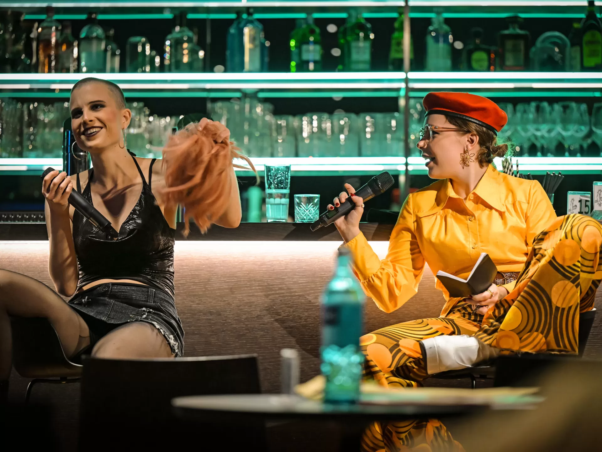 Lisa hält eine Perücke in der Hand und blickt lachend nach vorne. Noémie blickt Lisa von der Seite an und lacht.