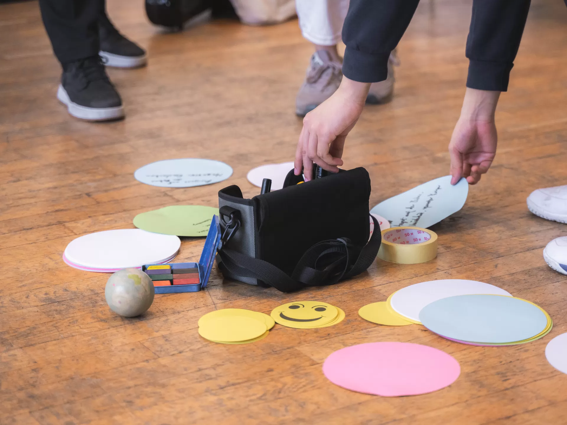 Zwei Hände greifen sich Zettel und Stifte, die in einer Tasche auf dem Boden stehen.