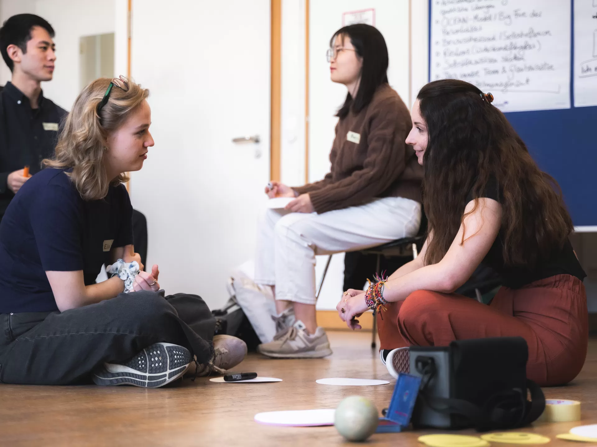 Zwei Studentinnen sitzen im Schneidersitz auf dem Boden eines Seminarraums und unterhalten sich. Um sie herum liegen Stifte und Zettel. Hinter ihnen sitzen zwei weitere Personen auf Stühlen und reden miteinander.
