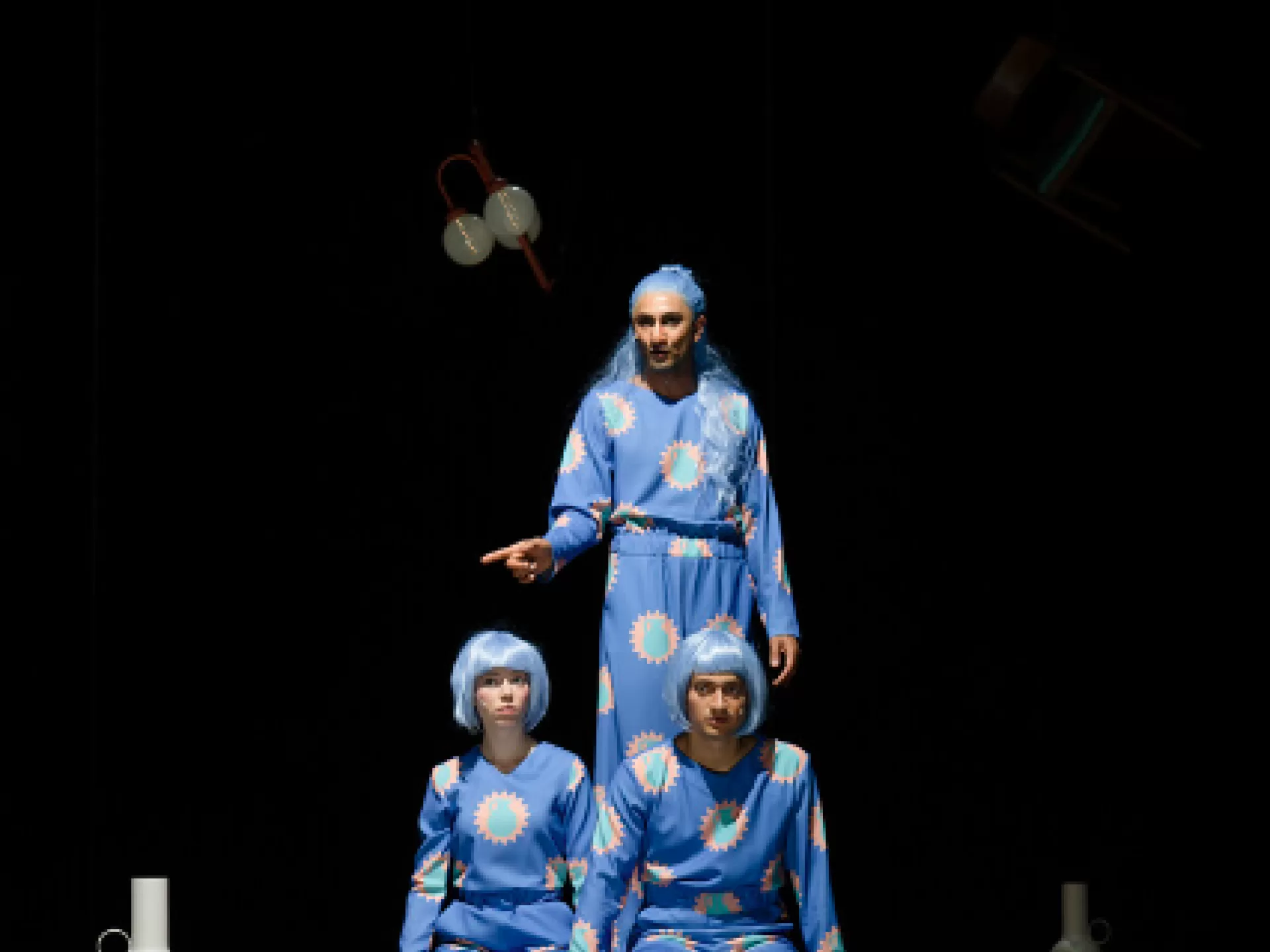 Zwei Menschen sitzen auf einer Kante vor einem dritten. Alle schauen nach rechts und tragen blaue Perücken und blaue Uniformen