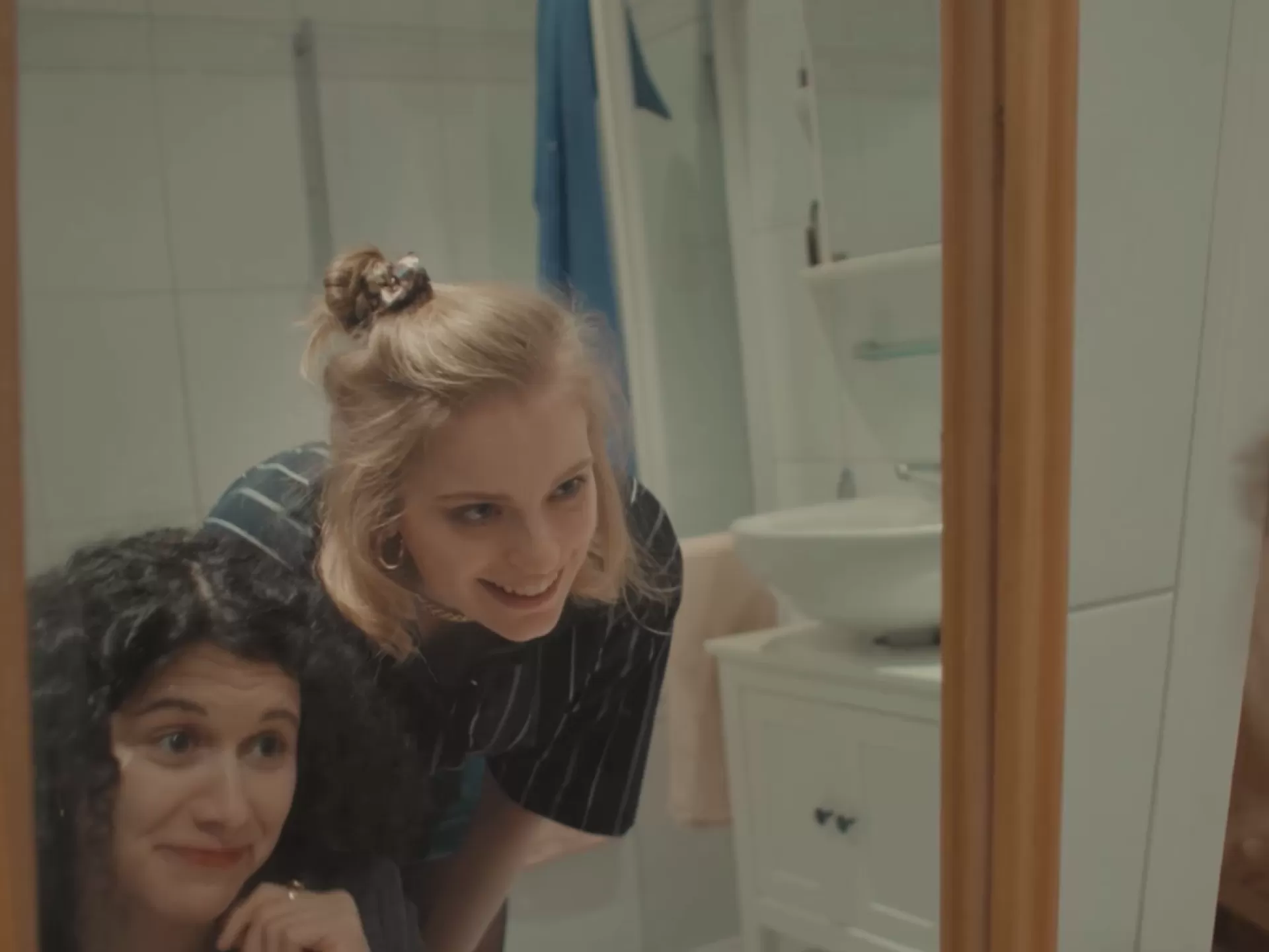 Zwei junge Frauen lächelnd vor einem großen Badezimmerspiegel.