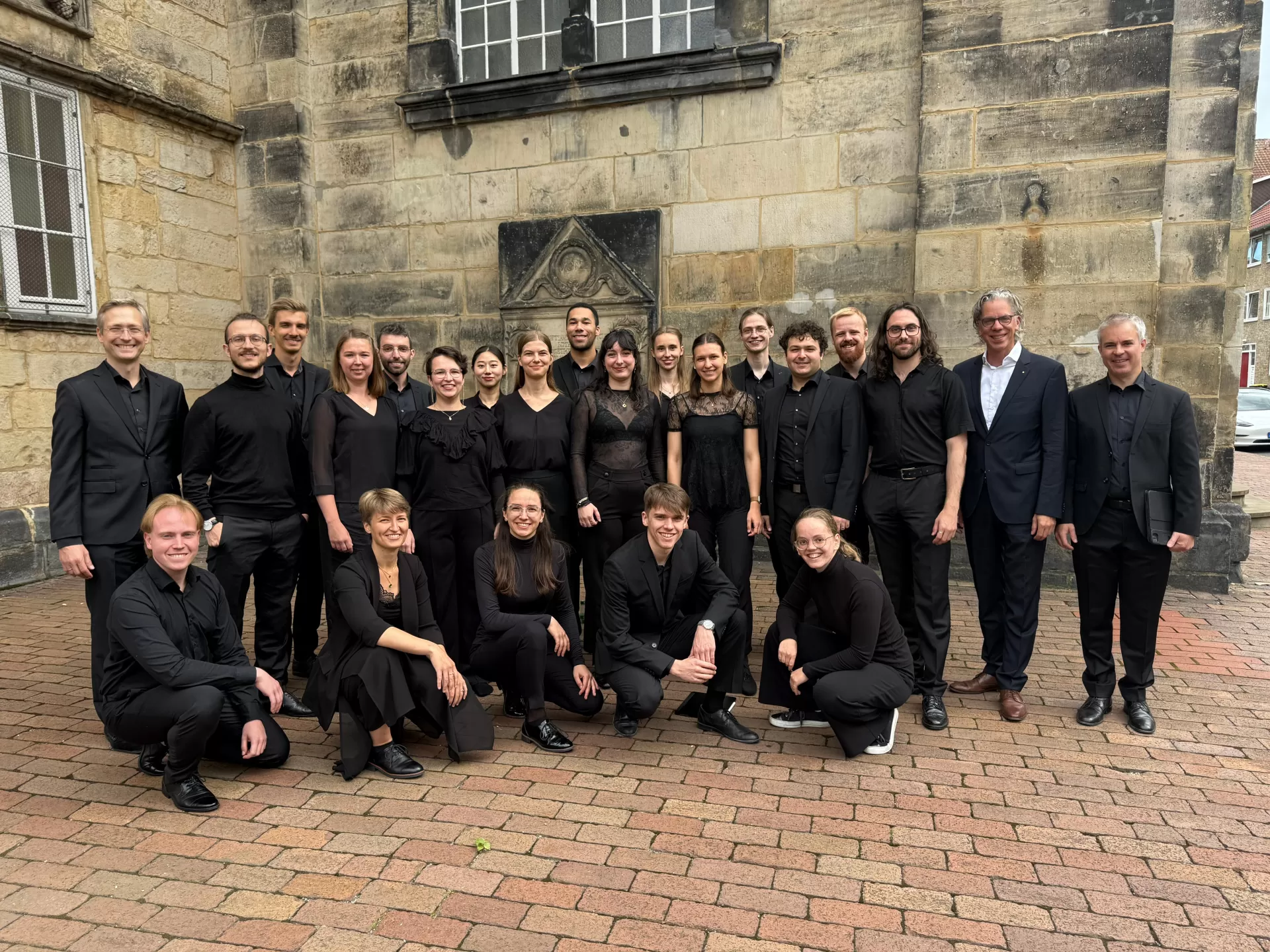 Gruppenfoto in mehreren Reihen, alle in schwarzer Konzertkleidung vor der Kirche