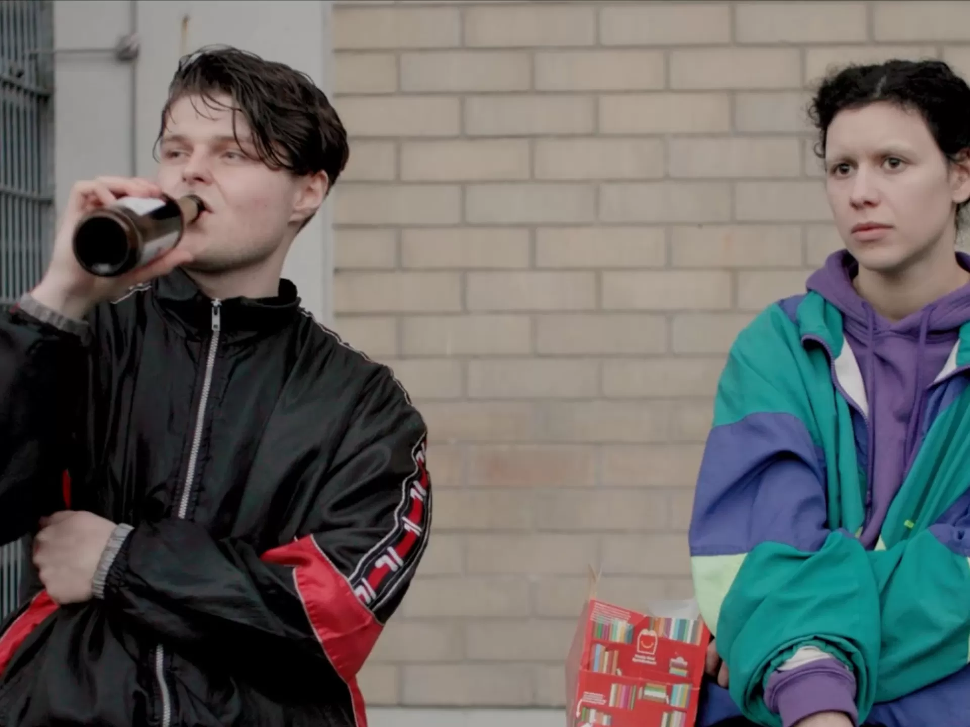 Zwei junge Personen in Jogginganzügen, einer trinkt ein Bier aus der Flasche.