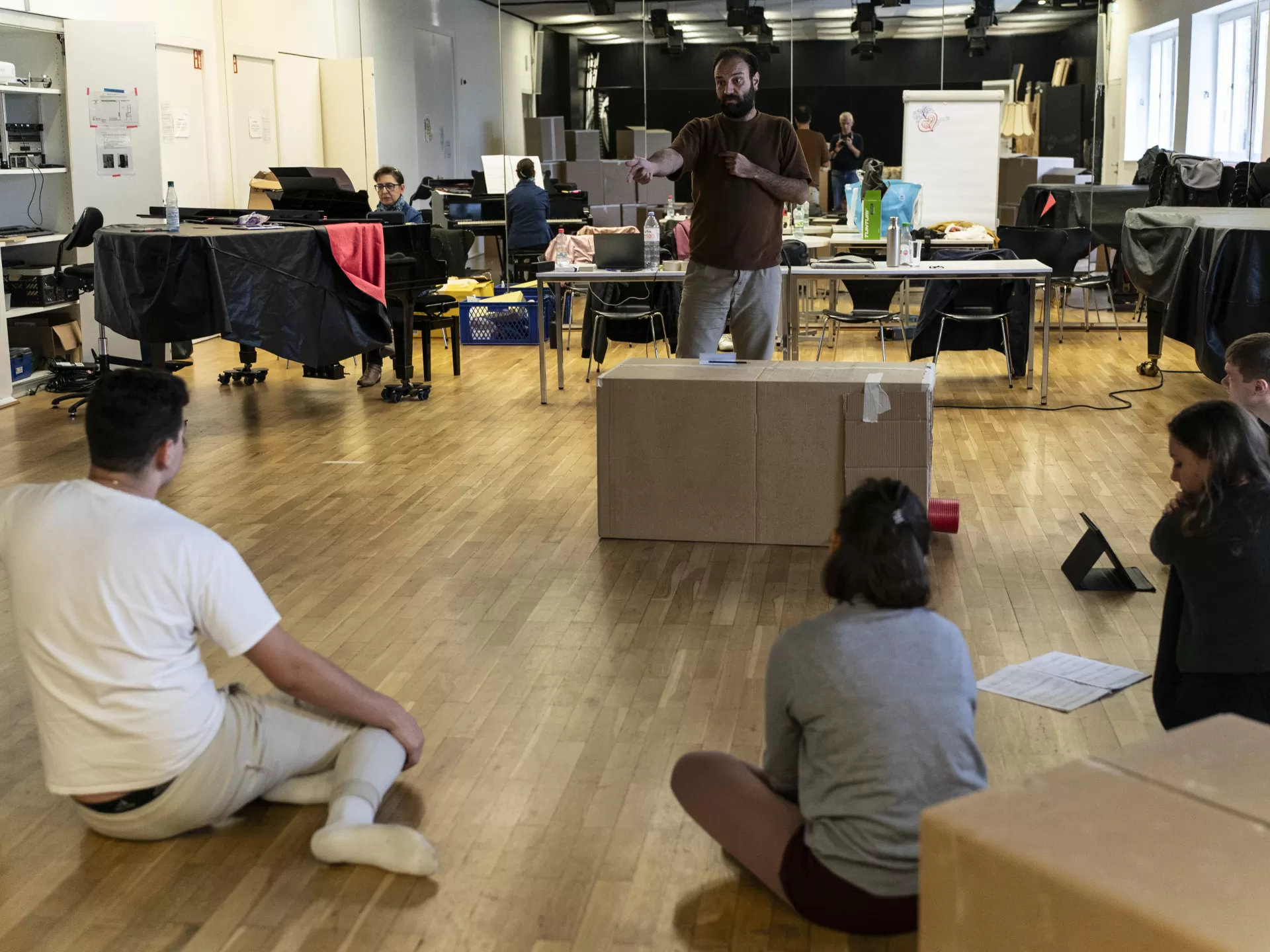 Proben im Opernstudio, ein Mann gibt Regieanweisungen, während die Darstellenden auf dem Boden sitzen