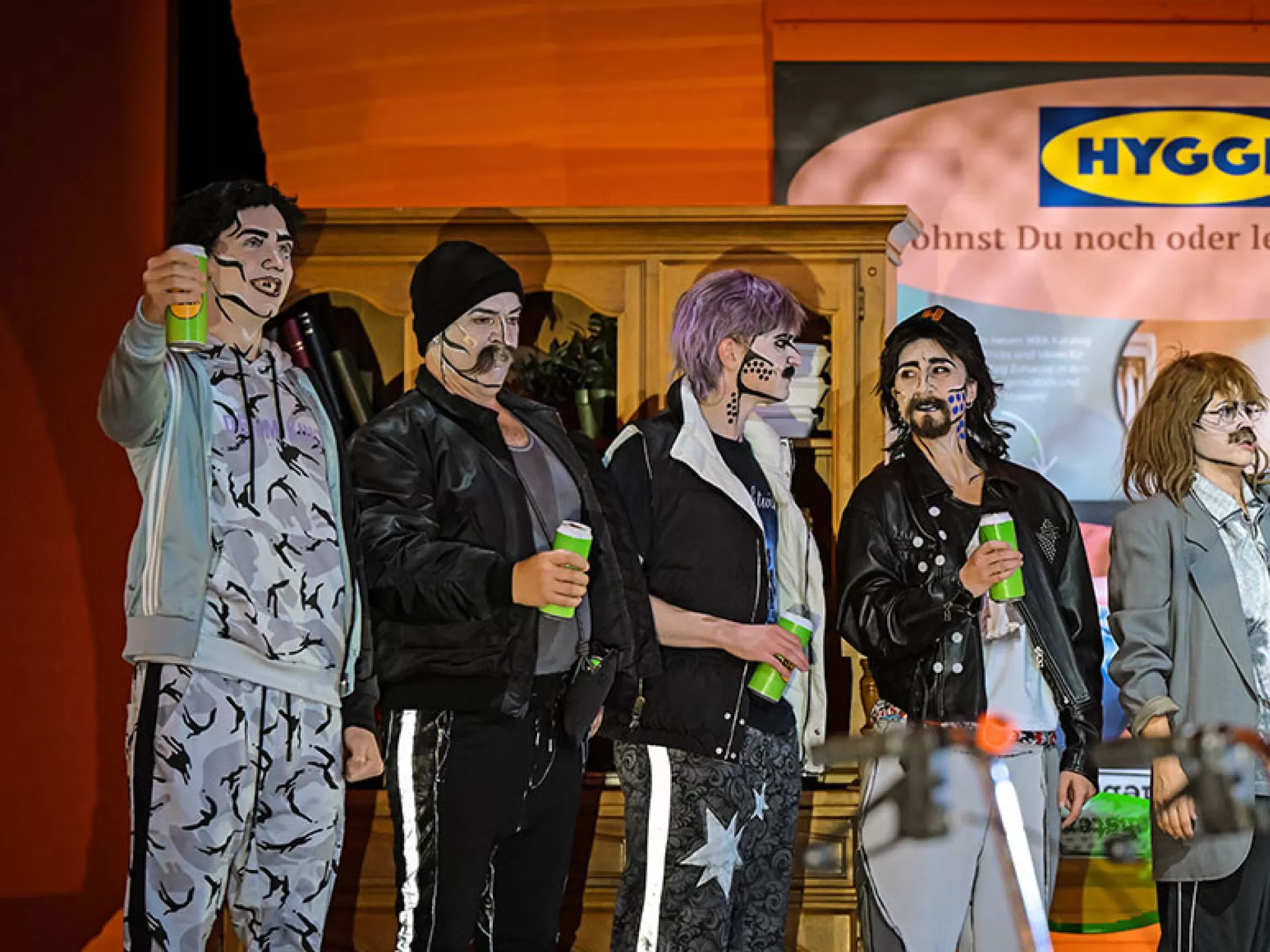 Eine große Personengruppe, alle tragen schwarze, weiße oder graue Klamotten. Sie sind im Pop Art Stil geschminkt. Der Hintergrund ist orange