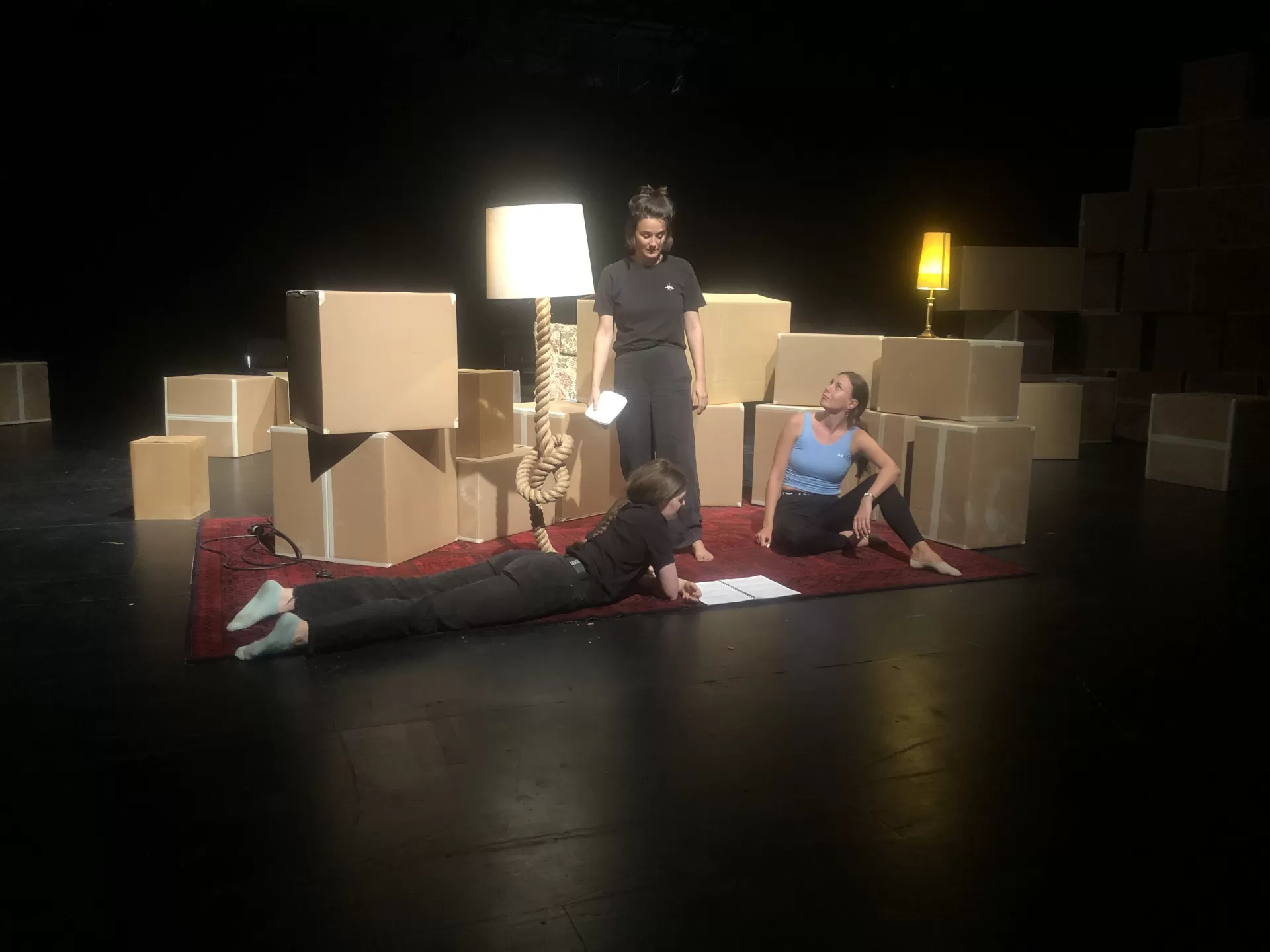 Szenenprobe auf der Bühne, drei Darstellerinnen in einem Bühnenbild aus Pappkartons, Lampen und einem Teppich