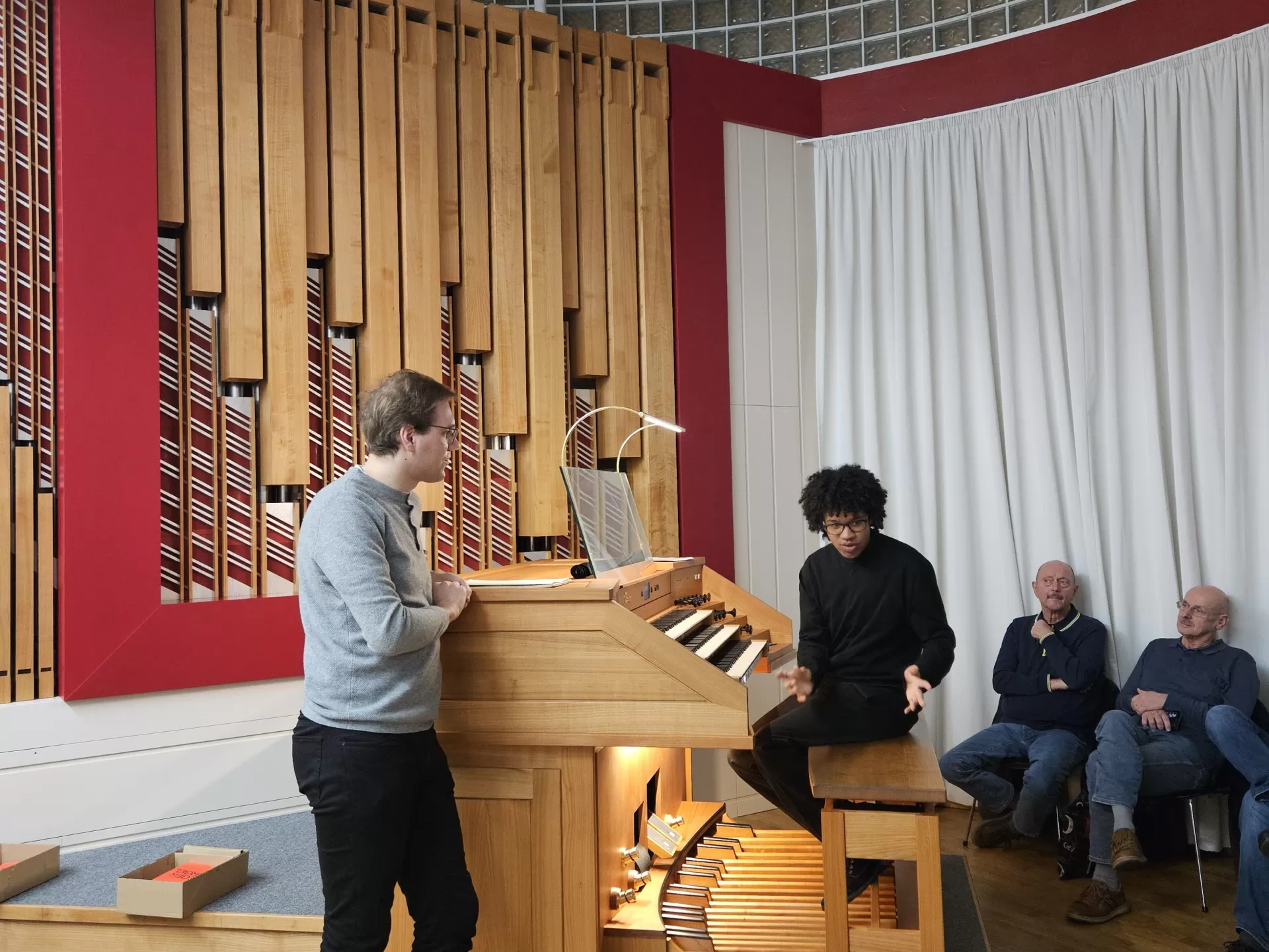 Zwei Studenten bei einer Orgelführung. Einer sitzt auf der Orgelbank und gestikuliert mit seinen Armen, der andere steht daneben. Im Raum sitzen mehrere Personen
