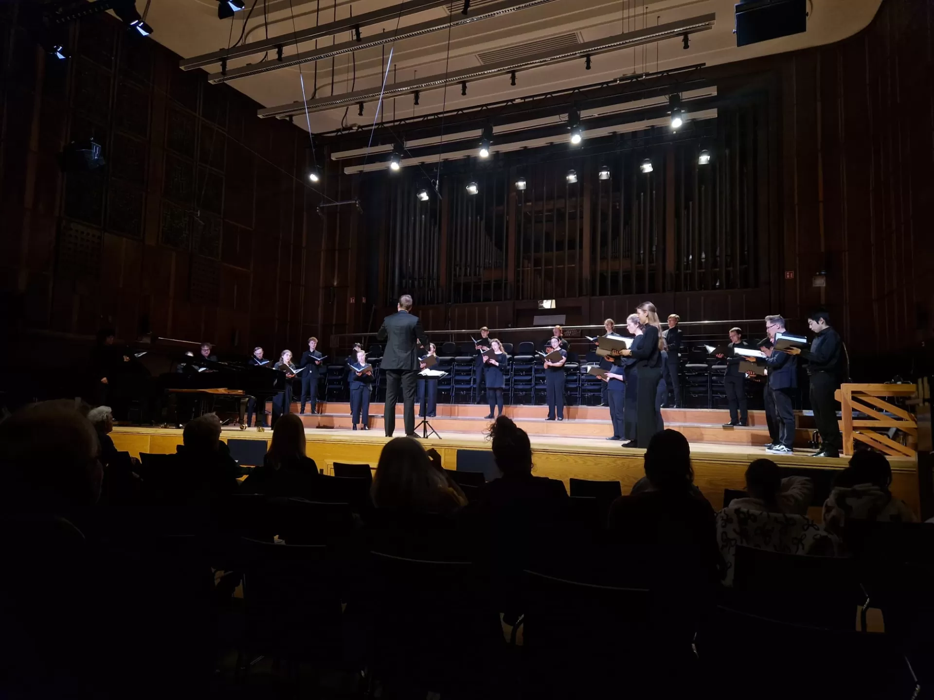 Blick in Großen Saal, der Chor steht mit etwas Abstand zueinander auf der Bühne und singt. Ein Student steht mittig und dirigiert.