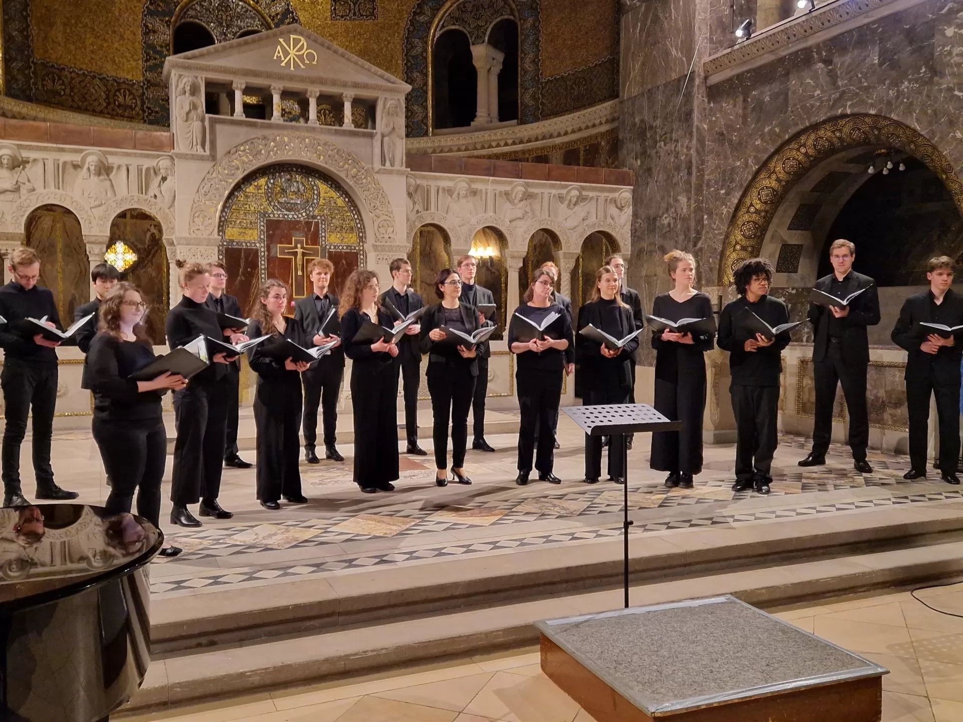 Die Mitglieder des StudioChors stehen mit offenen Noten in der Hand im Altarraum der Erlöserkirche, während Benjamin Hartmann vor ihnen steht und eine Begrüßungsansprache hält.