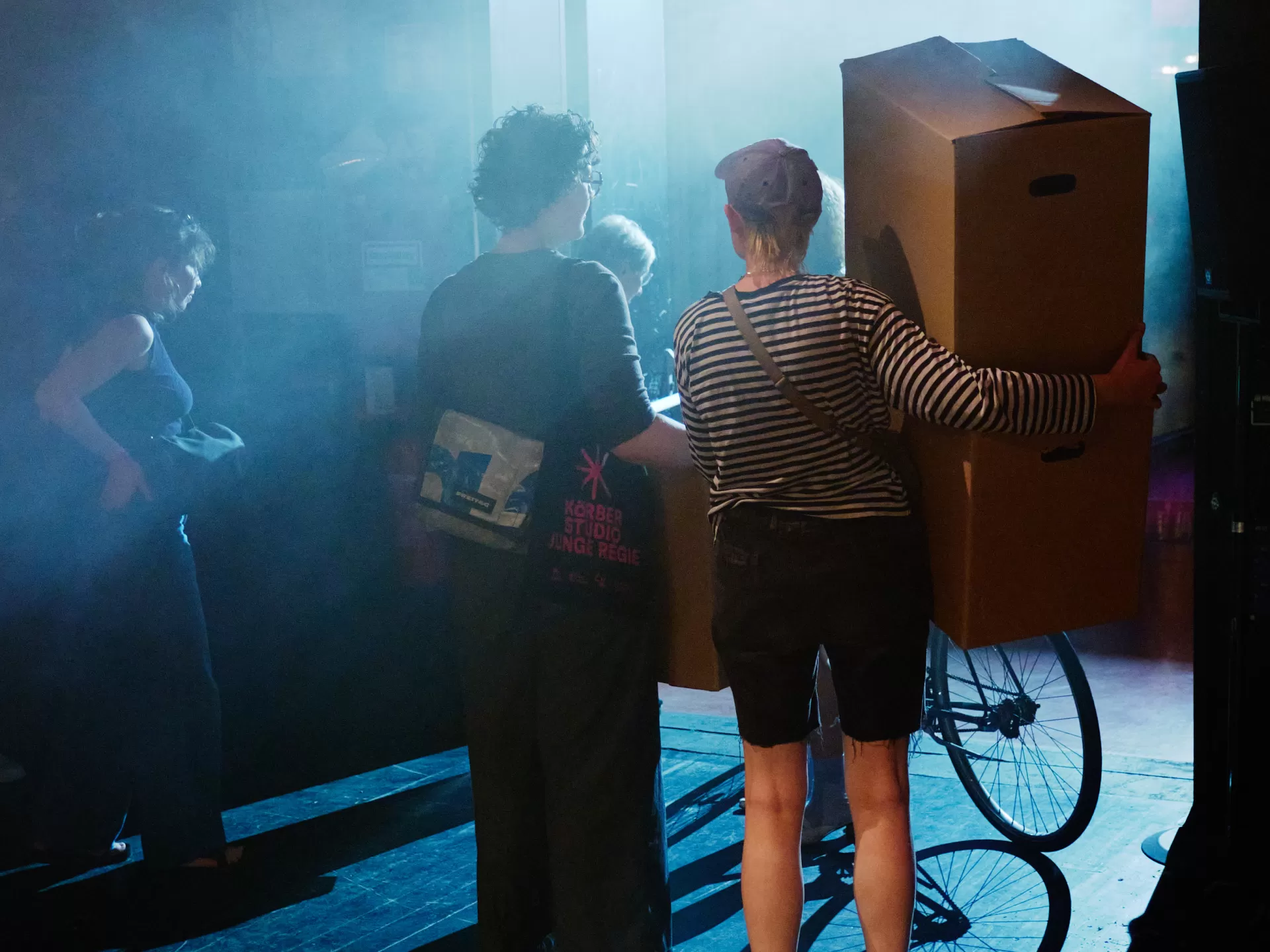 Im Vordergrund stehen zwei Frauen, sie sind von hinten zu sehen. Vor ihnen ein helles bläuliches Licht, die Schatten gehen in Richtung Kamera. Beide Frauen haben einen Umzugskarton in der Hand. Links neben ihnen und teils verdeckt vor ihnen stehen weitere Personen sowie ein Fahrrad. 