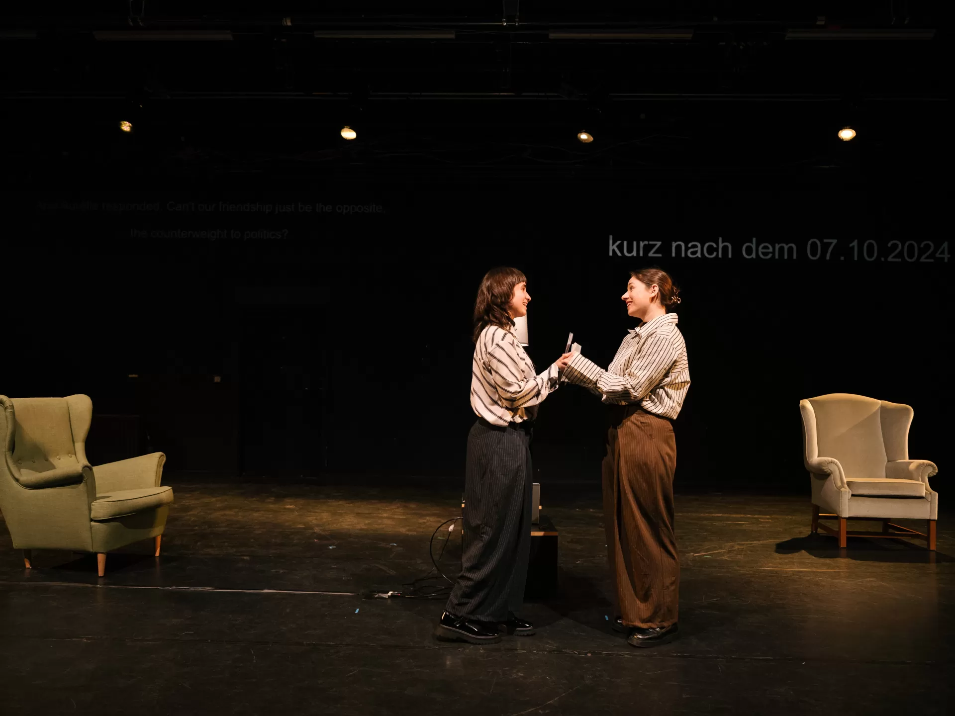 Zwei Sessel rechts und links im Hintergrund. Dazwischen stehen sich zwei junge Frauen lächelnd gegenüber und halten sich an den Händen