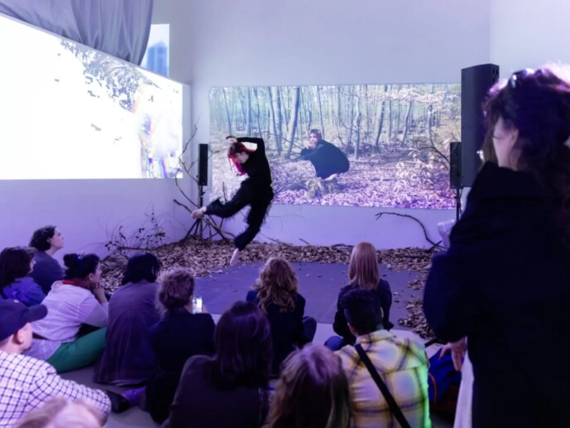 eine junge Frau in schwarz springt in die Luft hinter ihr eine Videoprojektion von ihr selbst im Wald