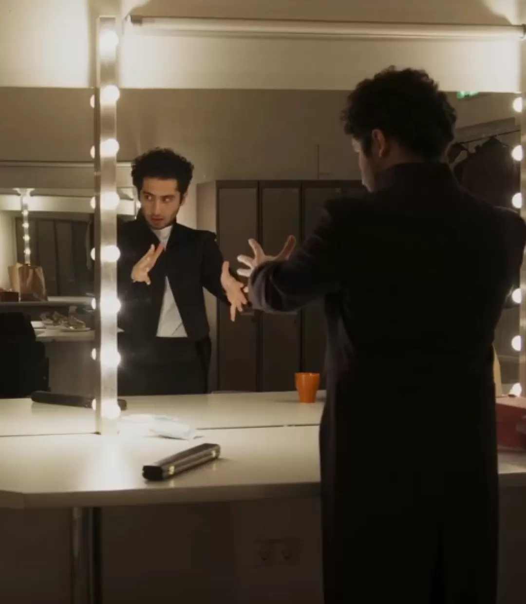 Ein Dirigent steht nervös und mit ausgestreckten Armen vor dem Spiegel in seiner Garderobe.