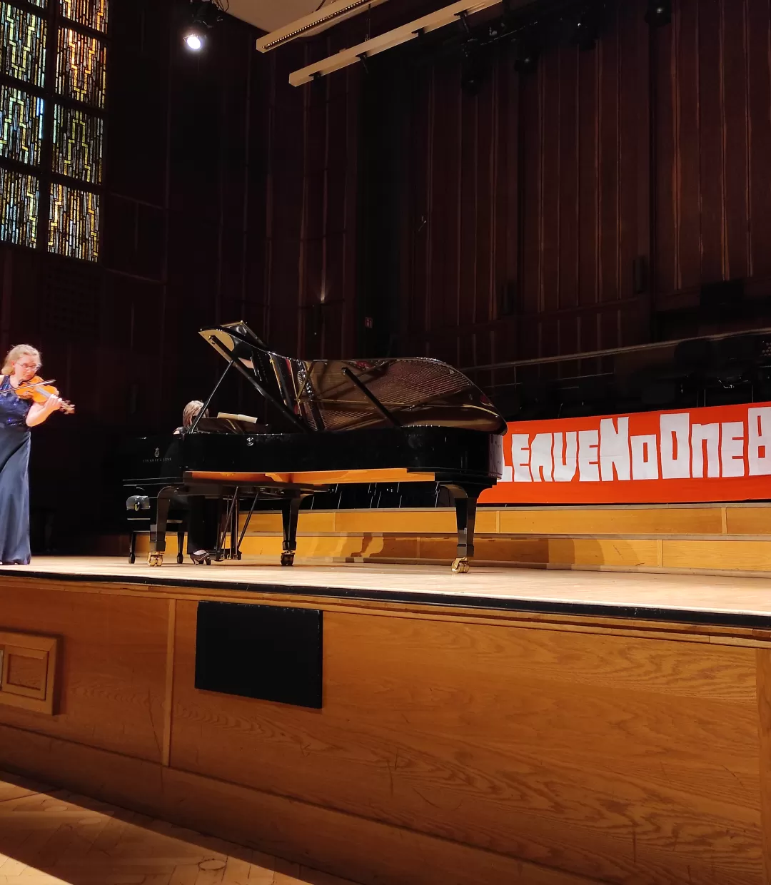 Konzert im Großen Saal der HfMDK. Auf der Bühne ein Flügel und eine Geigerin, hinten am Bühnenrand hängt ein Transparent mit der Aufschrift Leave No One Behind.