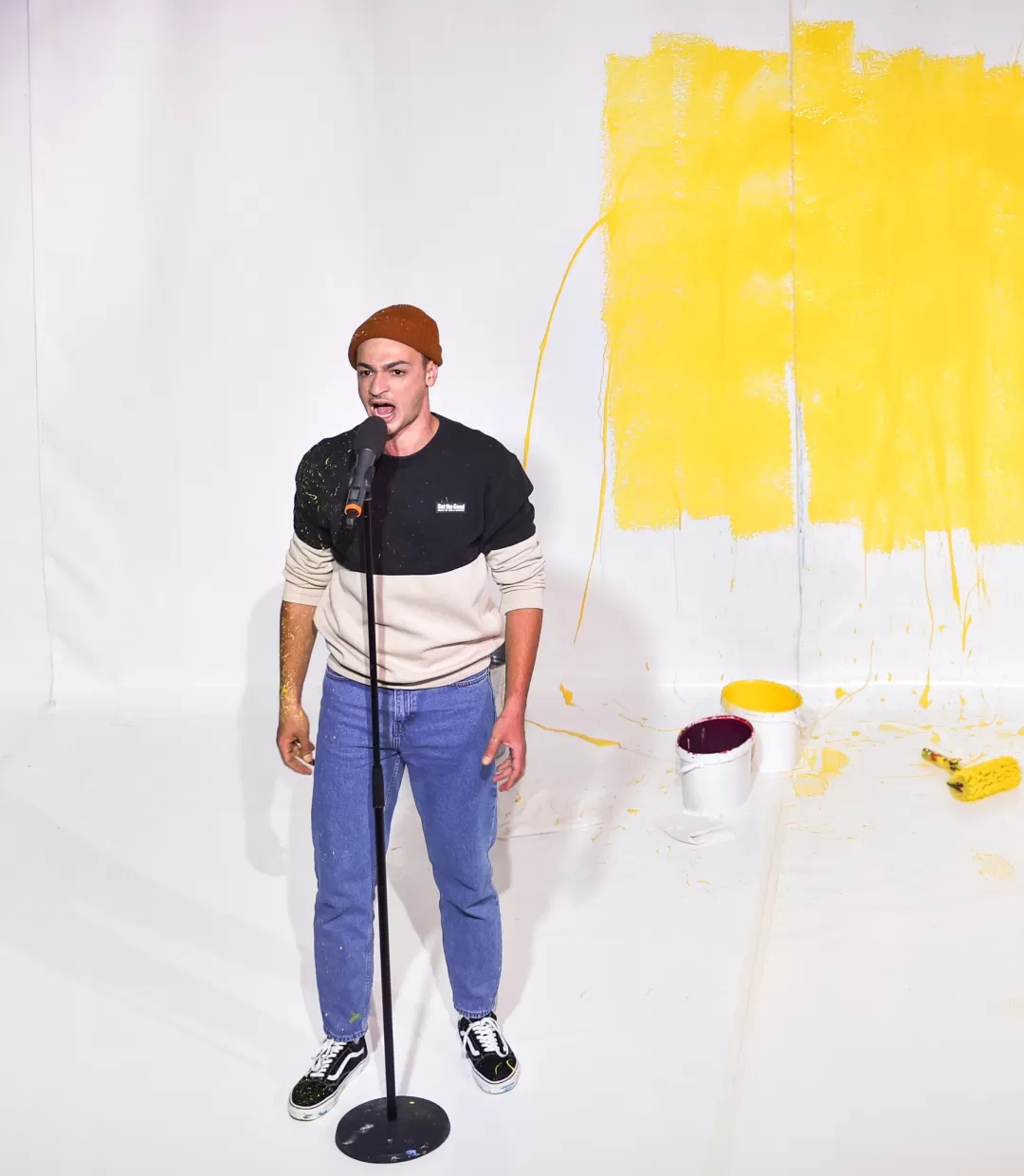 Schauspieler steht vor an einem Stehmikrofon vor einer weißen Wand, davor liegen Farbeimer und -rollen.