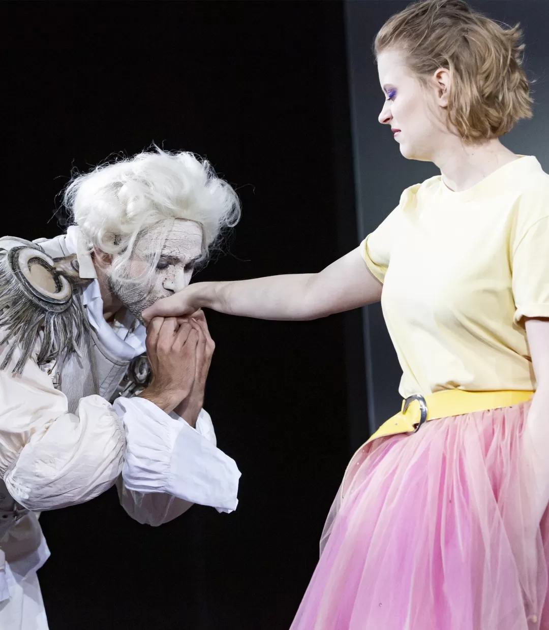 Schauspieler mit gepuderter Perücke gibt einer Schauspielerin, die angewidert schaut, einen Handkuss.