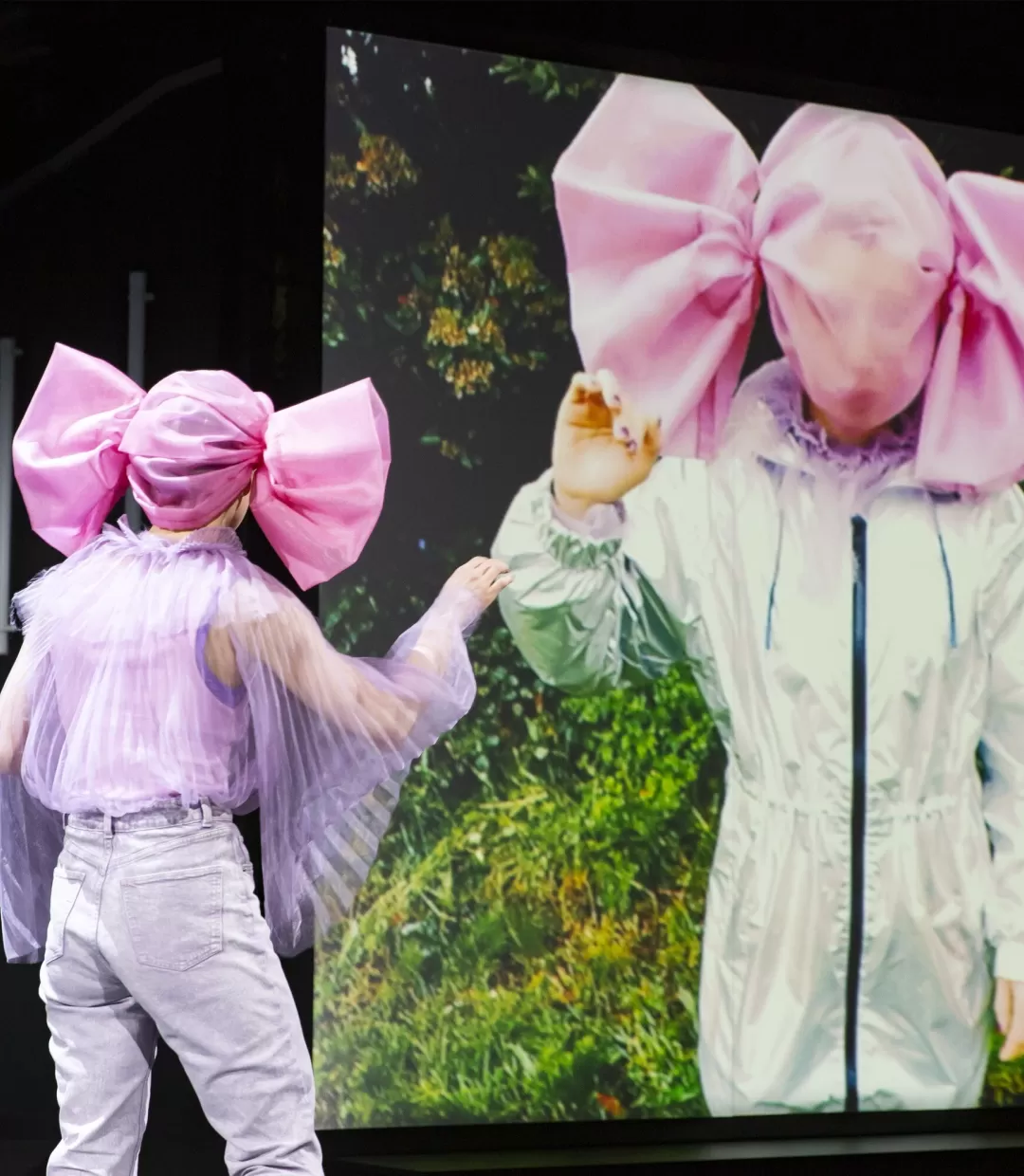 Schauspielerin mit einer Kopfbedeckung, die wie ein großes Bonbon aussieht, steht vor einer Videoleinwand.