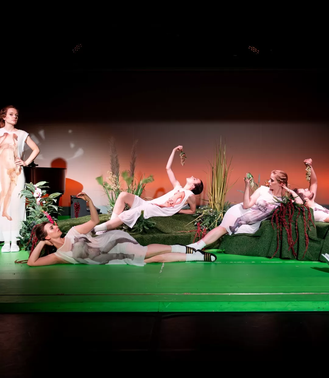 Gruppenfoto der Schauspieler*innen, stehend und liegend, Trauben essend.