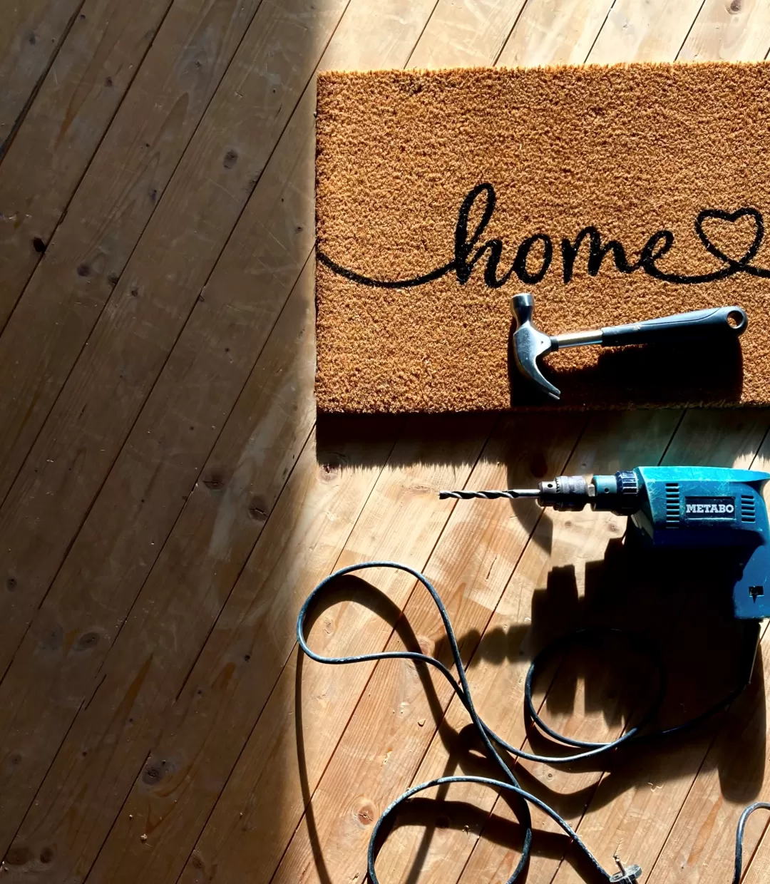 Aus der Vogelperspektive sieht man einen Holzboden, auf diesem rechts an der Seite eine Fußmatte mit dem Wort "Home", darunter rechts ein blauer Elektrobohrer mit Strohkabel.