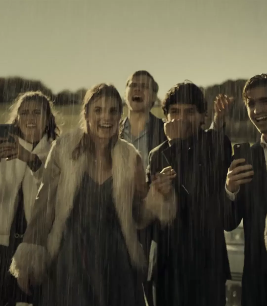 Gruppe im Regen stehend und lachend, machen mit ihren Handys Fotos von einem Polizisten.