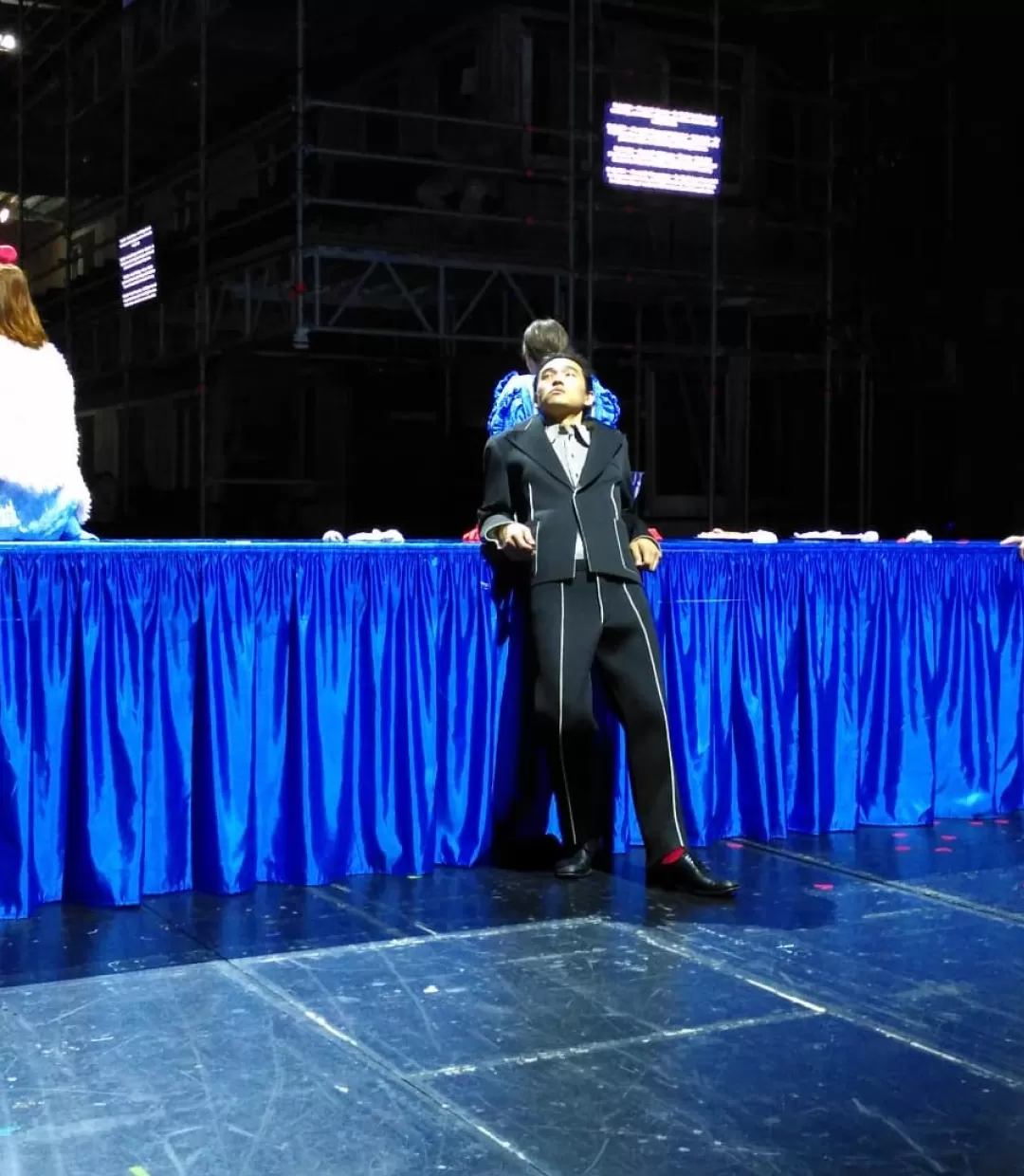 Auf einer Bühne steht ein ca. fünf Meter langer und ein Meter hoher schmaler Tisch, der von einer bodenlangen blauen Satindecke bedeckt ist. Links darauf sitzt eine Person, die von hinten zu sehen ist, mit schulterlangem rötlichem Haar und einer weißen Felljacke. In der Mitte vor dem Tisch steht ein Mann im schwarzen Anzug, er schaut gelangweilt nach rechts links oben. Direkt dahinter sitzt eine weitere Person auf dem Tisch mit dem Rücken zur Kamera. Weiter rechts vor dem Tisch steht ein zweiter Mann.