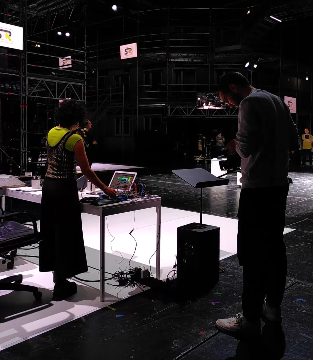 Im Vordergrund steht ein Tisch, darauf ein Laptop, Kabel, ein Aktenordner, davor ein Schreibtischstuhl neben dem eine Frau steht, die von hinten zu sehen ist. Rechts daneben ein Mann, der nach unten auf einen leeren Notenständer blickt. Weiter Hintergrund sind im Bereich einer Bühne weitere Personen zu erkennen. Es handelt sich um eine Probensituation.