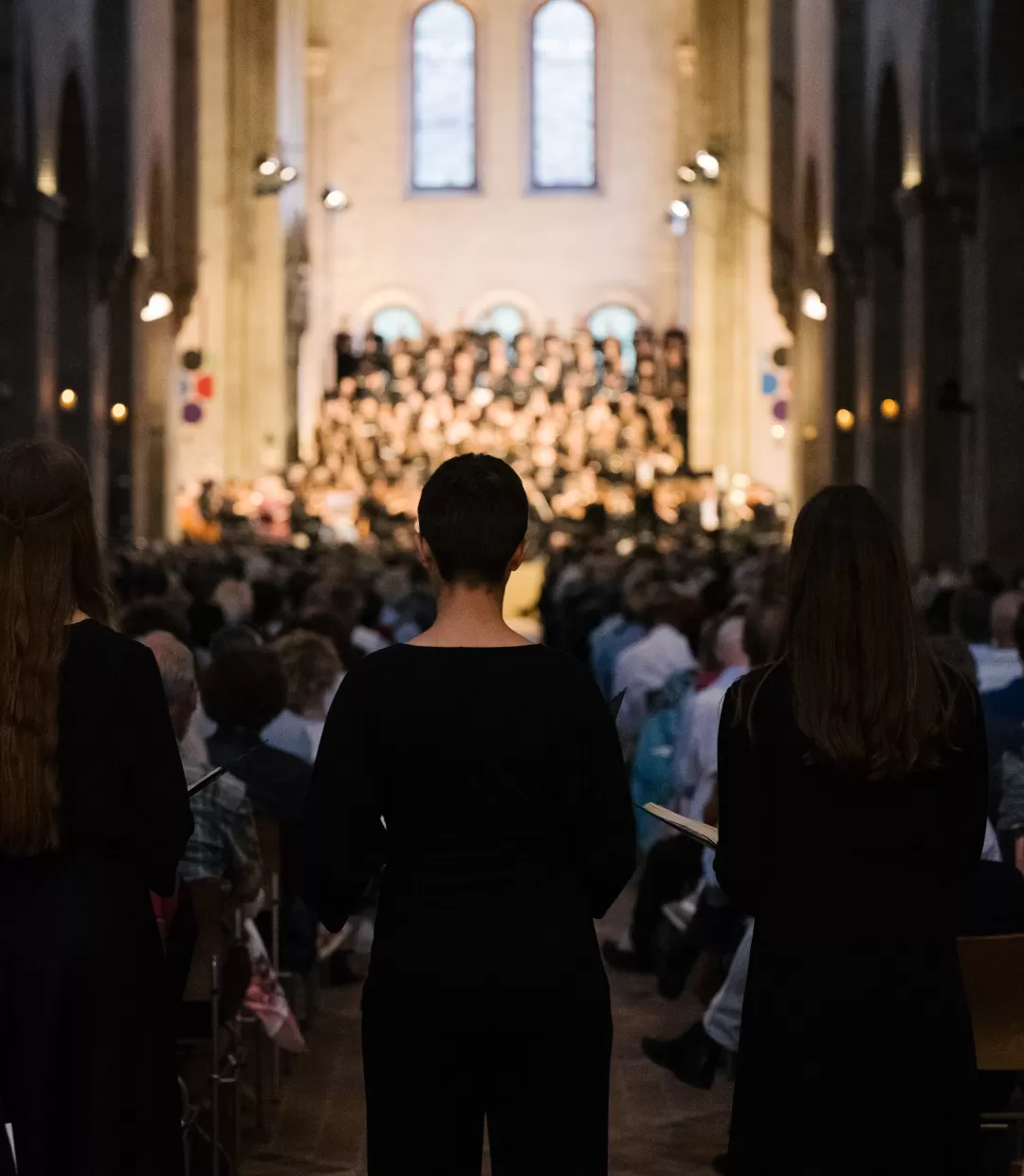 3 Studentinnen von hinten fotografiert, im Hintergrund sieht man verschwommen Chor und Orchester auf der Bühne im Kloster Eberbach