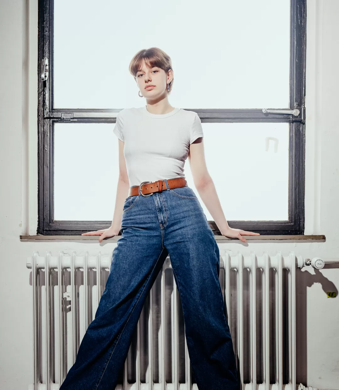Totale Paula Marie Schulze, blaue Jeans und ein weißes T-Shirt, im Hintergrund ein Fenster und ein weisser Heizkörper 