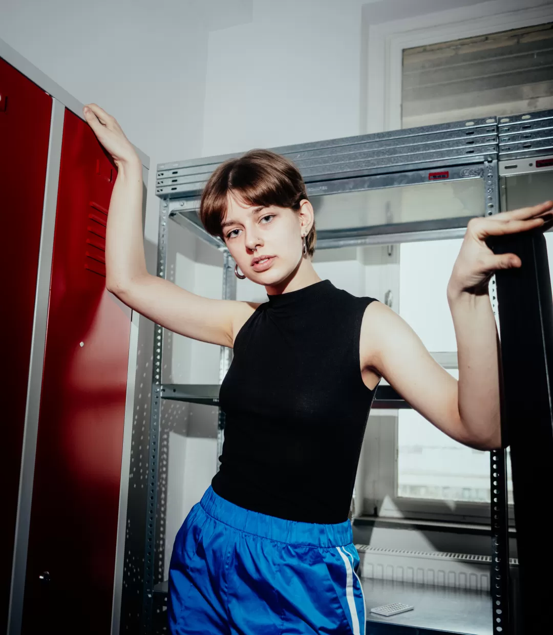 Halbtotale Paula Marie Schulze, blaue Hose, schwarzes Top, im Hintergrund ein roter Spind und ein graues Metallregal 
