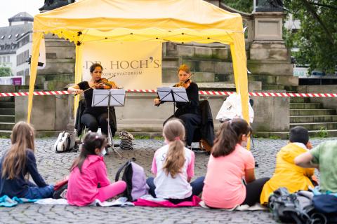 Zwei Musikerinnnen spielen unter einem gelben Pavillon, davor auf dem Boden sitzen mehrere Kinder.