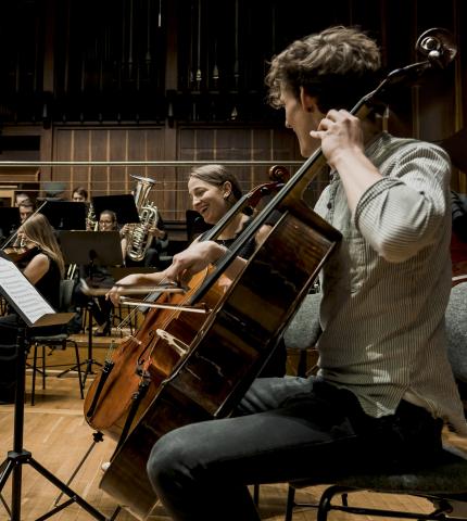 Eine Cellistin und ein Cellist in der Orchesterprobe, lachend einander zugewandt.