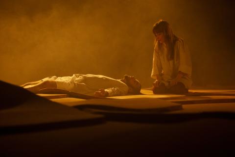 Ein Schauspieler liegt mit geschlossenen Augen auf dem Rücken. Eine Schauspielerin sitzt neben ihm und betrachtet ihn. Die Bühne in orangenem Licht und Nebel.