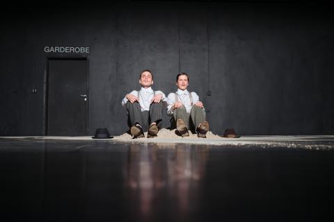 Zwei Männer sitzen nebeneinander am Boden in einem kleinen Sandhaufen vor einer schwarzen Wand. Jeder hat ein weisses Hemd und eine Hose mit Hosenträgern an, neben ihnen liegt jeweils ein Hut am Boden..