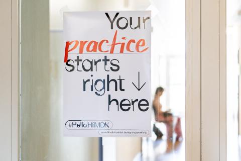 Plakat an einer Glastür, die zu Überäumen der HfMDK führt. Aufschrift: Your practice starts right here.
