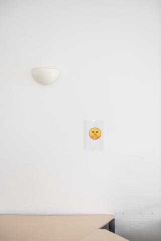 Ein Zettel mit einem gelben Emoji mit Finger auf dem Mund hängt an einer weißen Wand