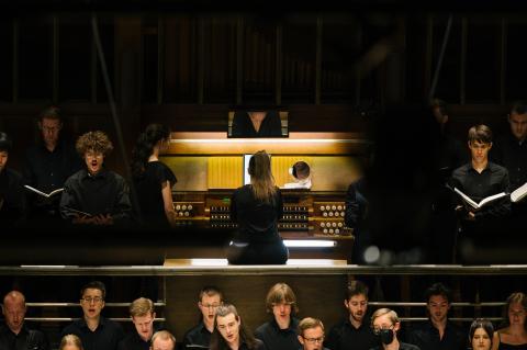Organistin an der Orgel im Großen Saal der HfMDK, davor und daneben Chorsänger*innen.
