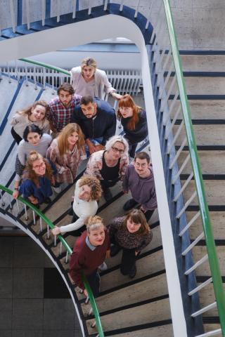 Die Studierenden und die Studiengangleitung des Studiengangs BA Regie stehen auf einer geschwungenen Treppe und schauen nach oben in die Kamera.