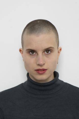 Das Bild zeigt einen Headshot von Lisa Freiberger. Sie isst dezent geschminkt, trägt die Haare in einem Buzzcut und hat einen dunkelgrünen Rollkragenpullover an. 