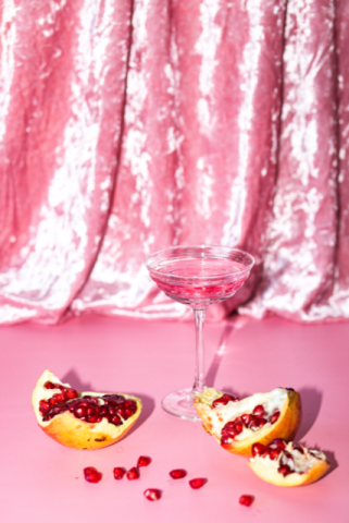 Ein Glas mit einem Drink vor einem pinken Vorhang. Davor liegt ein aufgebrochener Granatapfel.